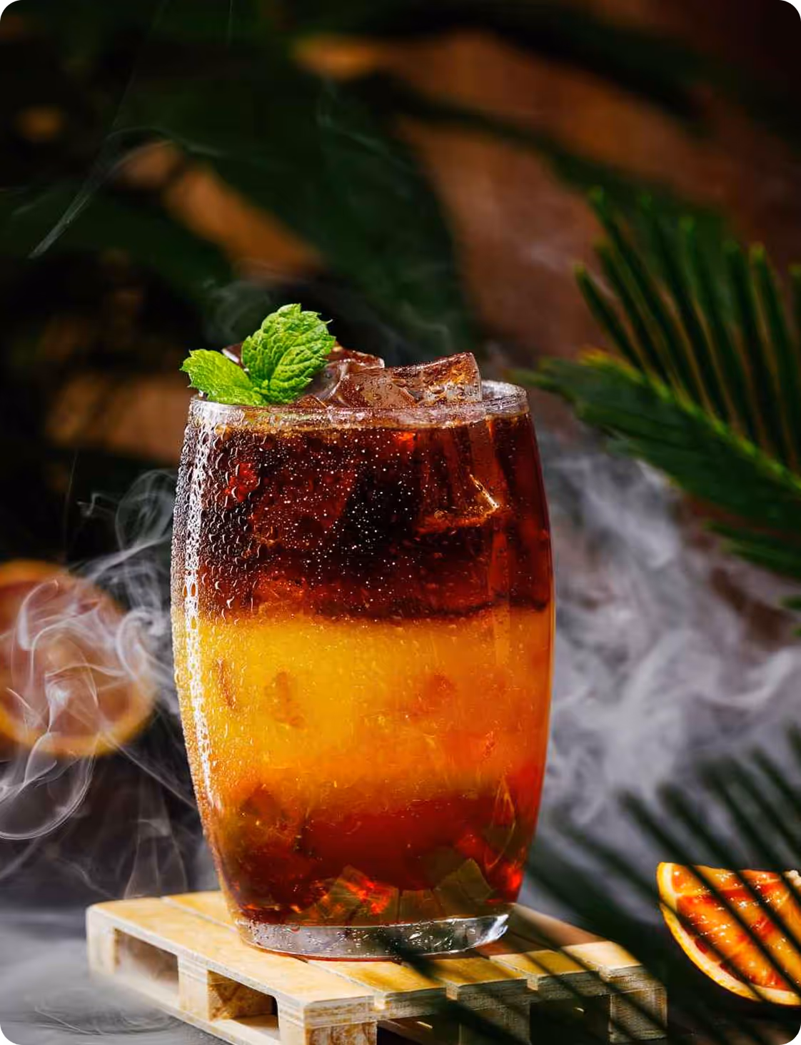 Glass of iced layered cola and orange juice garnished with mint on a wooden coaster with smoky background.